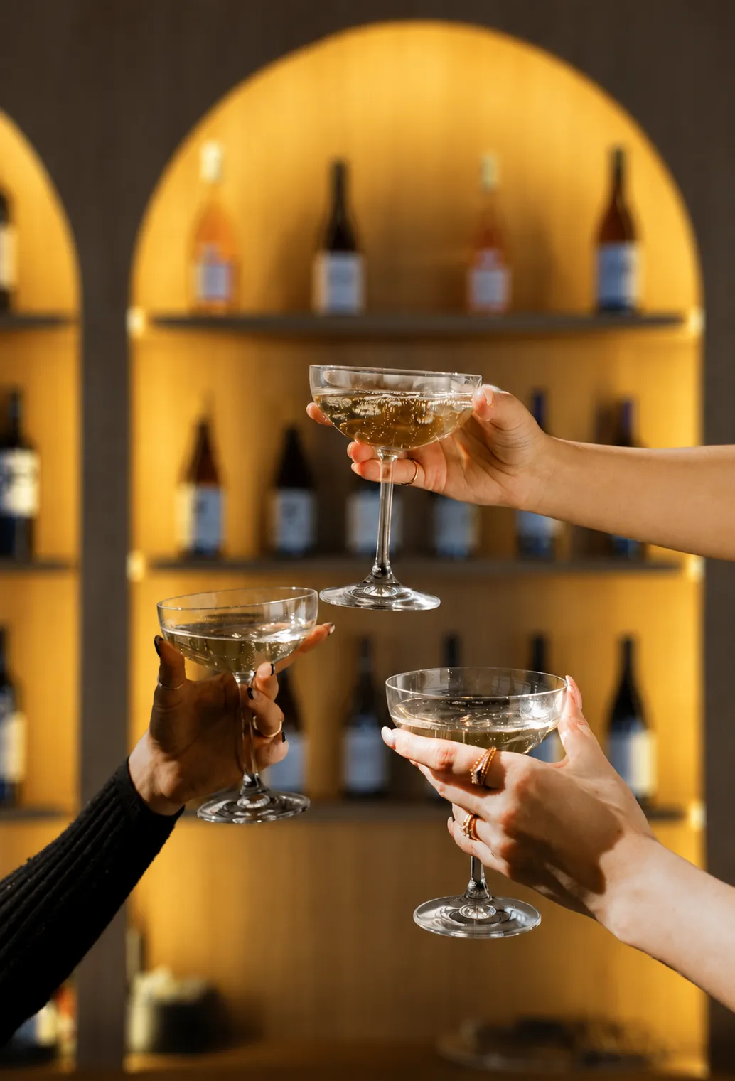 Tres manos brindando con copas de cava frente a la vinoteca iluminada